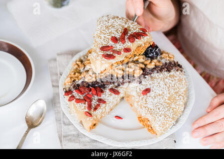 Woman's hands holding a piece of raw vegan sweet carrot cake with cream topped with walnuts, goji berries, chocolate truffles, coconut flakes and coco Stock Photo