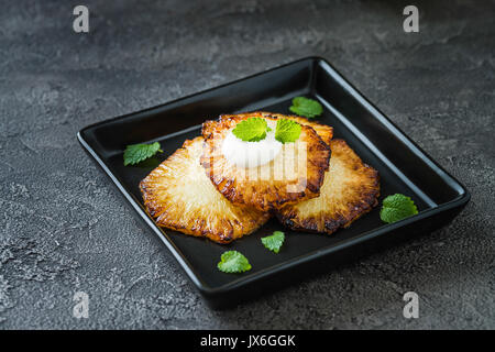 Fried pineapple slices with creme fraiche, mint in black square plate on dark background Stock Photo