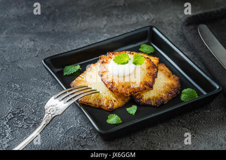 Fried pineapple slices with creme fraiche, mint in black square plate on dark background Stock Photo