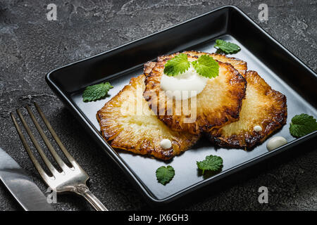 Fried pineapple slices with creme fraiche, mint in black square plate on dark background Stock Photo