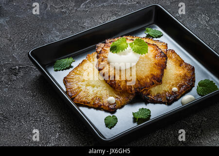 Fried pineapple slices with creme fraiche, mint in black square plate on dark background Stock Photo