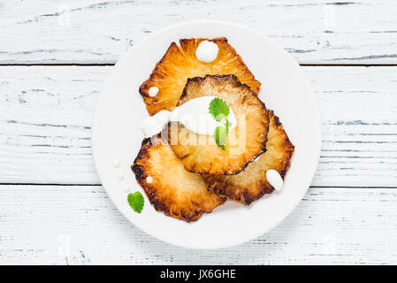Fried pineapple slices with creme fraiche, mint in white plate on wooden background. Top view Stock Photo