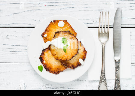 Fried pineapple slices with creme fraiche, mint in white plate on wooden background. Top view Stock Photo