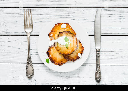 Fried pineapple slices with creme fraiche, mint in white plate on wooden background. Top view Stock Photo