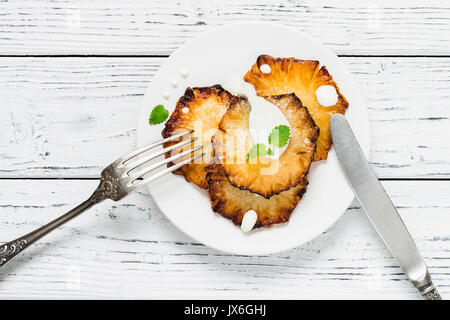 Fried pineapple slices with creme fraiche, mint in white plate on wooden background. Top view Stock Photo
