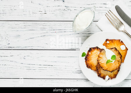 Fried pineapple slices with creme fraiche, mint in white plate on wooden background. Top view, copy space Stock Photo