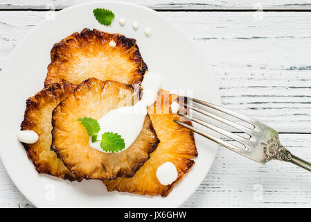 Fried pineapple slices with creme fraiche, mint in white plate on wooden background. Top view Stock Photo