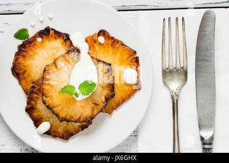 Fried pineapple slices with creme fraiche, mint in white plate on wooden background. Top view Stock Photo