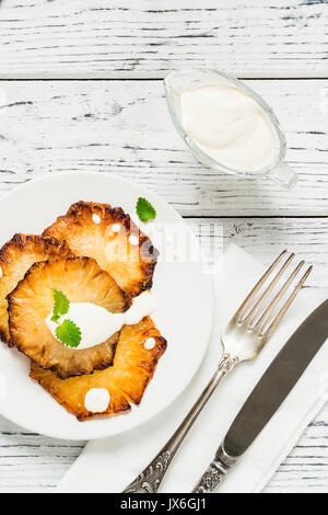 Fried pineapple slices with creme fraiche, mint in white plate on wooden background. Top view Stock Photo