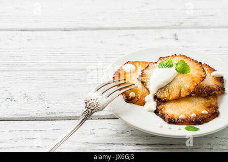 Fried pineapple slices with creme fraiche, mint in white plate on wooden background. Copy space Stock Photo