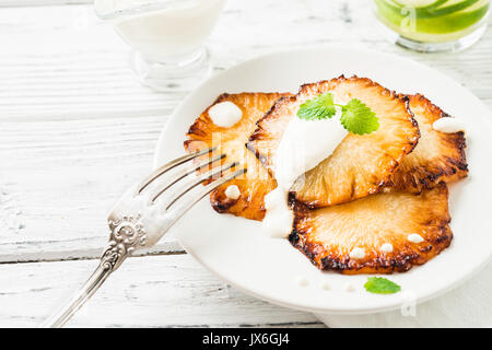 Fried pineapple slices with creme fraiche, mint in white plate on wooden background Stock Photo