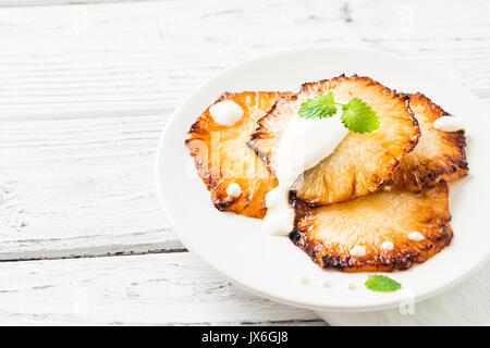 Fried pineapple slices with creme fraiche, mint in white plate on wooden background. Copy space Stock Photo