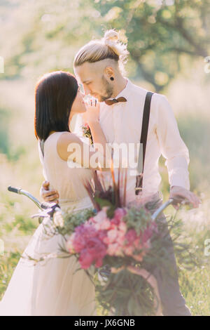 The close-up vertical photo of the lovely newlyweds carrying the bicycle with the bouquet of pink flowers at the background of the sunny forest. Stock Photo