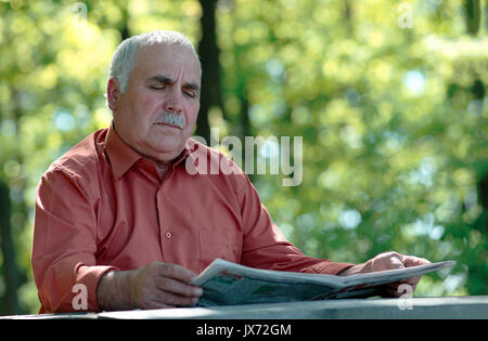 Senior man sitting reading a newspaper at a rustic wooden table in a wooded garden relaxing as he catches up on the latest news Stock Photo