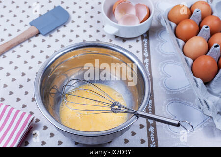 Beaten eggs and a hand whisk in a stainless steel bowl on a country kitcken table with a box of eggs Stock Photo