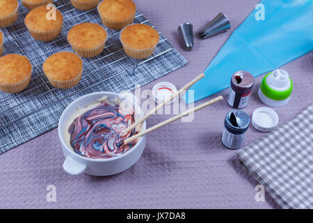 Fresh homemade cupcakes on a baking tray with a bowl of multi coloured buttercream ready to be piped onto the cakes Stock Photo