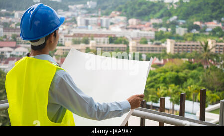 A construction engineer. Wearing a helmet and vest holding a plans with blur city or building background in construction concept Stock Photo