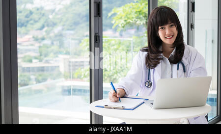 Young asia doctor are Smiling and writing a report for a treatment plan with Stethoscope,The hand holding the pen and find information with laptops in Stock Photo