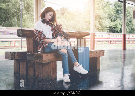 Traveler girl using tablet with a luggage sitting and waiting for train on the station. Outdoor adventure travel by train concept with vintage filter Stock Photo