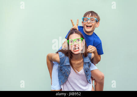 Funny emotions and feelings. The freckled brother climbed up on the back of a older cute sister. Making funny crazy face and looking at camera. Indoor Stock Photo