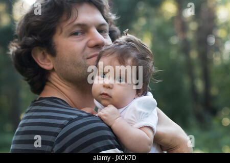 Dad is holding a daughter in his arms for a walk in the park. Spring day, family walk in nature, sunny. Focus on the baby. Soft focus. Stock Photo