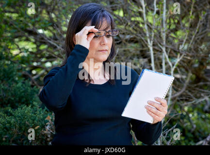 Attractive middle aged woman wearing reading glasses, looking down at notebook in her hands in an outdoor setting Stock Photo