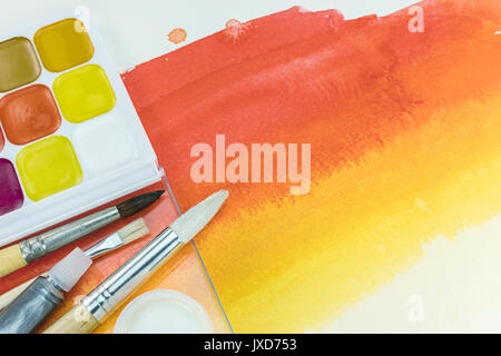 palette of watercolor paints and various paintbrushes on red yellow watercolor background Stock Photo