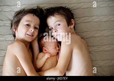 Two children, toddler and his big brother, hugging and kissing their newborn baby brother at home, few days after delivery Stock Photo