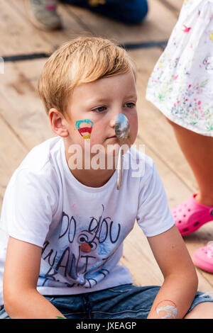 England, Broadstairs folk week. Hobby Horse club. Blonde Child, boy, 8-9 years old, sitting on wooden deck, face painted with colourful heart and a spoon hanging from his nose. Stock Photo