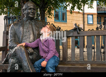 Close up young boy with glasses admiring bronze commemorative sculpture of Sandy Irvine Robertson, local wine merchant on a bench in Leith, Edinburgh Stock Photo