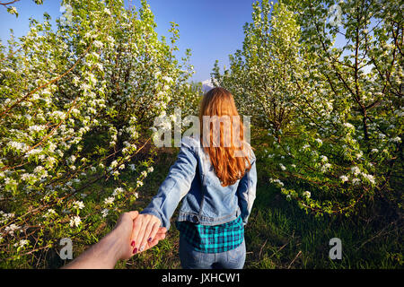 Woman in with red hair holding her friend by hand and leading him to the garden with cherry blossom trees at sunrise Stock Photo