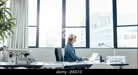 Side view of female executive sitting in her office using laptop. Young caucasian woman looking busy working on laptop. Stock Photo
