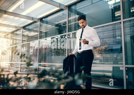Handsome business traveler looking at his smart phone outside airport. Businessman standing with luggage and using smart phone. Stock Photo