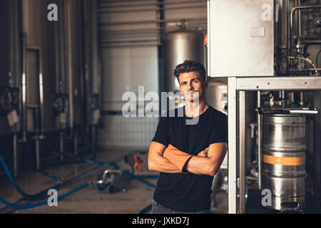 Portrait of young man standing by beer filling machine. Brewery worker with industrial equipment. Stock Photo