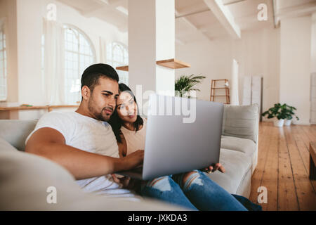 Loving young couple sitting on sofa and using laptop computer. Young man and woman relaxing on couch with laptop at home. Stock Photo