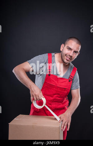 Mover guy packing box with duct tape and smiling on black background Stock Photo