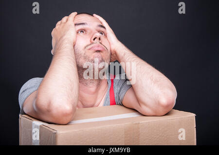 Mover guy making problem gesture on cardboard box with hands holding head on black background Stock Photo
