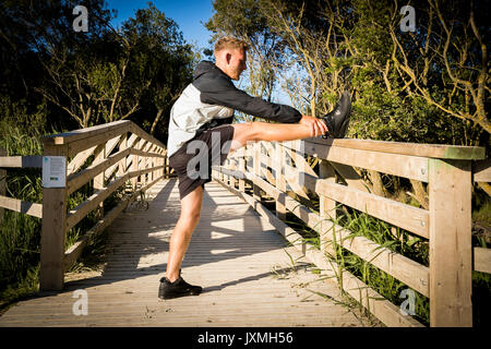 Young male runner warming up, stretching leg on rural footbridge Stock Photo