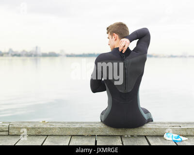 Rear view of man in wetsuit sitting on pier, Melbourne, Victoria, Australia, Oceania Stock Photo