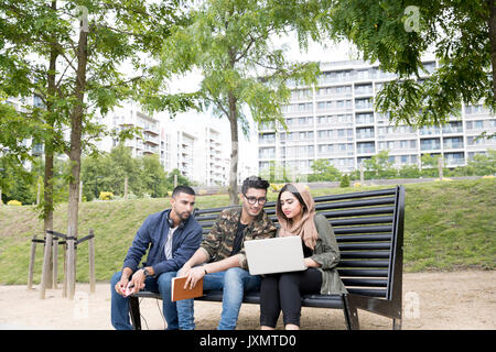 Three friends, sitting on bench in park, looking at laptop Stock Photo