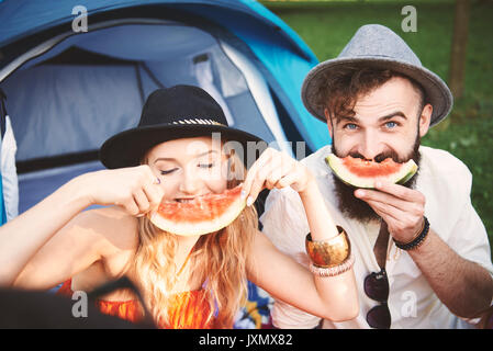 Young couple in trilbies making smiley face with melon slice at festival Stock Photo