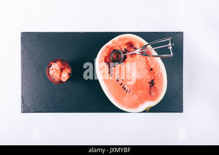 Half a ripe watermelon, slices in a glass and an ice cream spoon on a white table top view Stock Photo