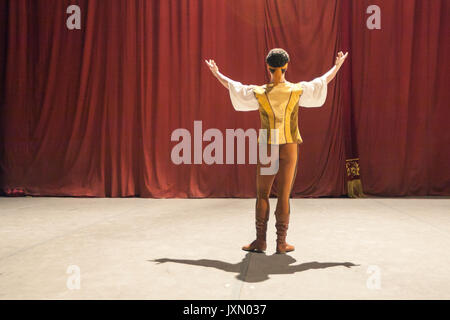 theater, other side, professionalism concept. on backstage ballet dancer in honey yellow vest, white shirt, pants and dancing boots, making a gesture of greeting to red curtains hiding auditorium Stock Photo