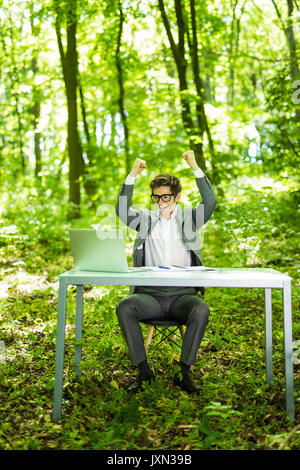 Portrait of handsome succesful business man in suit working at laptop at office table with raised hands in green forest park. Stock Photo