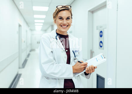 Portrait of happy young female doctor standing in hospital corridor and writing medical report on clipboard. Caucasian woman working in nursing home. Stock Photo