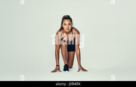 Confident female athlete in starting position ready for running. Young woman about to start a sprint over grey background. Stock Photo