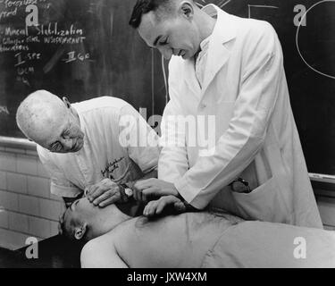 William Bennett Kouwenhoven, Candid photograph, Bending over man on table while demonstrating techniques of CPR, Cardiopulmonary resuscitation, with assistant in white coat in the foreground and blackboard in the background, 1969. Stock Photo