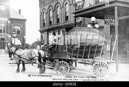 A racially prejudiced depiction of an African American man, who drives an open horse-carried wagon, placing his hand on an over-sized watermelon, which lies in the bed of the wagon on a street in front of a hotel in an American town, with the caption 'One of the melons, Watermelon Day, Webster City, [Iowa]', Europe, 1914. Stock Photo