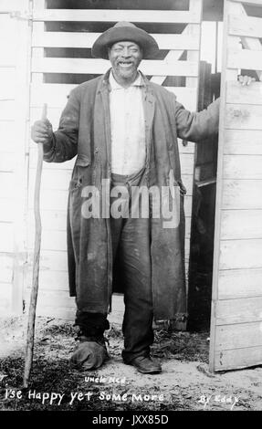 Full length standing portrait of a smiling mature African American man standing in front of a set of doors, carrying a walking stick, wearing a hat and a torn coat, with the racially prejudiced caption 'I'se Happy yet some more', 1915. Stock Photo