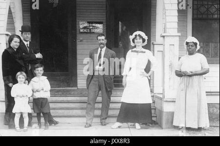 A family (consisting of a mature woman, a mature man, a young girl, and a young boy) and a boarding house staff (consisting of a mature man and two mature women) stand in front of the steps to a boarding house, 1915. Stock Photo